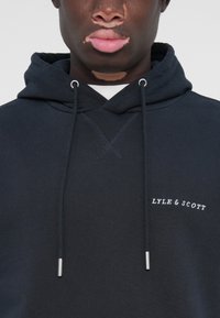 Marinblå hettegenser laget av mykt stoff, med en brodert kryssdesign, metall-aglets på snorene, og hvit "LYLE & SCOTT" logo.