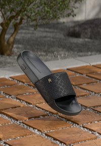 Sandale noire à bride ornée de motifs et petit logo doré, présentée au-dessus d'un sol en carreaux de bois et de galets avec un fond d'arbre flou.