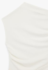 Primer plano de una camiseta blanca sin mangas con un cuello alto y un frunce sutil en los lados sobre una tela suave.