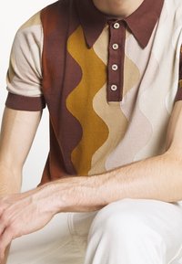 Muž oblečený v polo tričku s krátkým rukávem, které má vlnité vertikální pruhy v hnědé, hořčičné, béžové a růžové barvě, doplněné bílými kalhotami, sedí s rukou podepřenou o koleno.