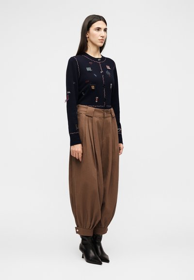 Γυναίκα με μακριά σκούρα μαλλιά που φοράει μια ναυτική ζακέτα με πολύχρωμες επιθέσεις και φαρδιά, ψηλόμεσα καφέ παντελόνια με γυρισμένα μπατζάκια στους αστραγάλους και μαύρα μπότες.