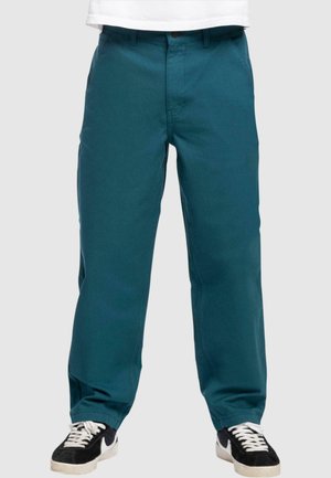 Türkisblaue Baumwollhose mit geradem Schnitt, mit Knopfverschluss, Gürtelschlaufen, Seitentaschen und glatter Textur; kombiniert mit schwarzen Sneakers.