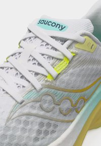 Λευκό παπούτσι για τρέξιμο Saucony με επάνω μέρος από δίχτυ, λευκά κορδόνια, λεπτομέρειες σε μέντα και κίτρινο, και λογότυπο της μάρκας στη γλώσσα και στο πλάι.