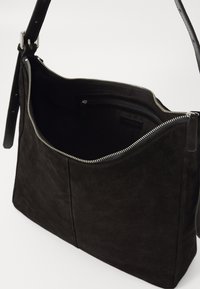 Borsa a spalla in suede nera con zip argentata, parzialmente aperta per mostrare il ampio scomparto principale e la tasca interna con zip.