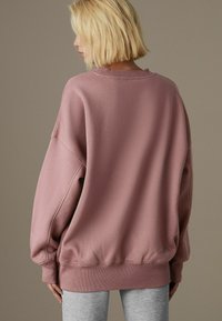 Przeszerzony różowy sweter wyświetlony od tyłu, z okrągłym dekoltem, prążkowanymi mankietami i miękką, teksturowaną tkaniną. Szare legginsy uzupełniają stylizację.
