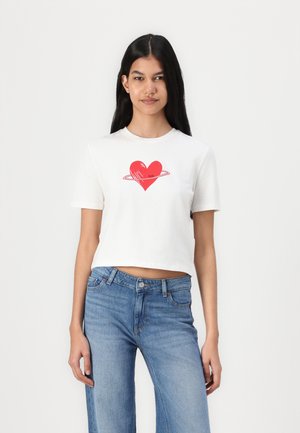 T-shirt bianco cropped con un design a cuore rosso accompagnato da un anello in orbita. Abbinato a jeans di denim blu. Texture di cotone liscia.