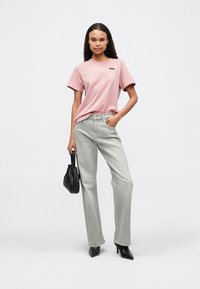 Γυναίκα στέκεται φορώντας ροζ μπλουζάκι, ανοιχτό γκρίζα τζιν, μαύρες γωνιαστές μπότες και κρατώντας μια μικρή μαύρη τσάντα.