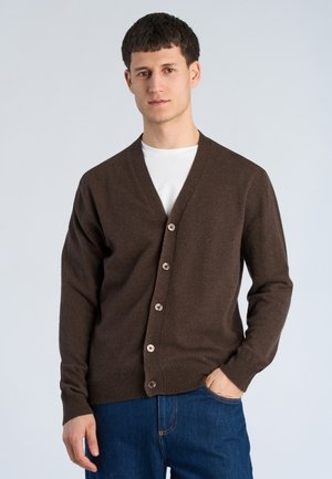Junger Mann trägt eine braune, geknöpfte Strickjacke über einem weißen Hemd und blauen Jeans, steht mit einer Hand in der Tasche vor einem einfarbigen Hintergrund.