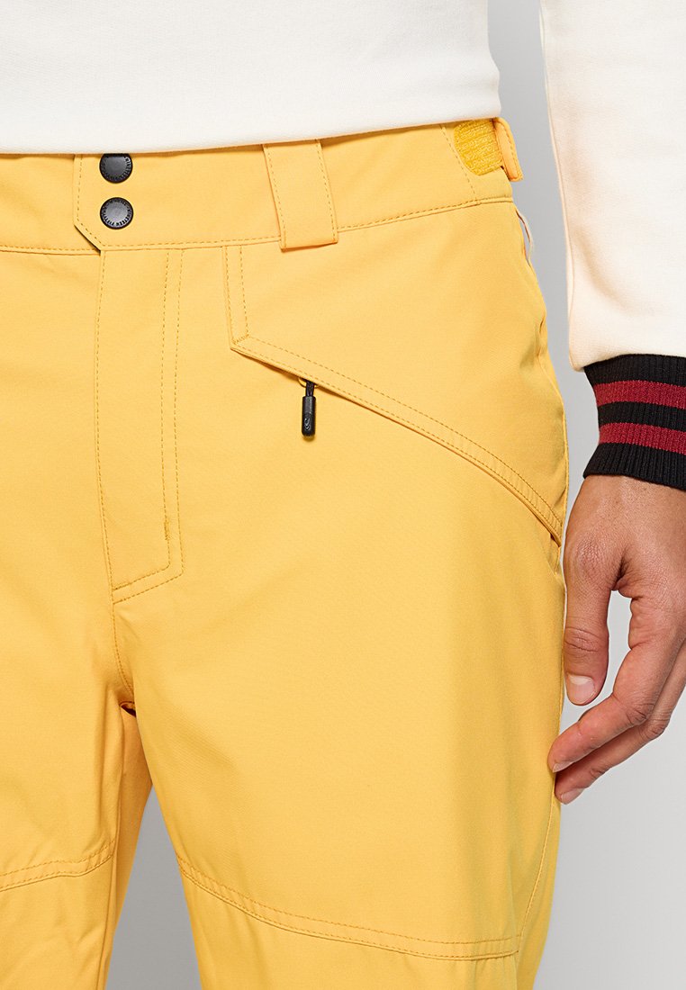 Gelbe Outdoor-Hosen mit einer Reißverschlusstasche, schwarzen Hardware-Details und einem strukturierten Stoff. Verfügen über gestichelte Nähte und einen Knopfverschluss.