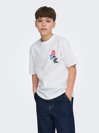Jongen met kort bruin haar, draagt een wit T-shirt met een rood-blauw bloemenprint en donkerblauwe spijkerbroek, handen in de zakken, neutrale achtergrond.