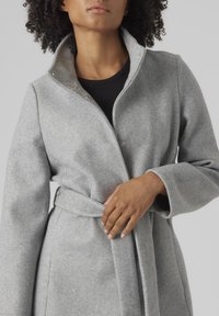 Manteau en mélange de laine gris avec un col haut, des fermetures à pression et une ceinture à nouer. Il a des manches longues et une texture douce pour la chaleur.