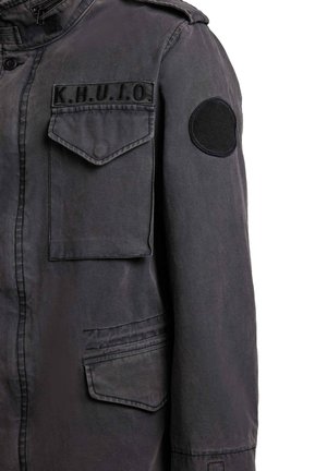 Mørkegrå jakke med brystlomme merket "K.H.U.J.O." og rund svart lapp på øvre erme, med klafflukkinger og synlige sømmer.