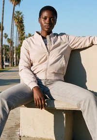 Fiatal személy világos dzsekiben és farmerben egy kültéri betonpadon ül, háttérben pálmafák és járda láthatók naplementekor.