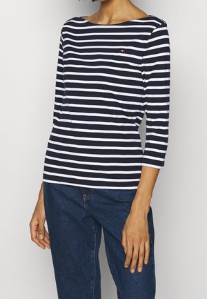 Kobieta ubrana w granatowo-biały, poziomo w paski, długi rękaw oraz ciemnoniebieskie dżinsy, stojąca na jednolitym tle.