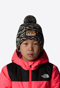 Czarna i biała czapka z dzianiny w wzór zebry, z kolorową naszywką tygrysa; pompon na górze. Noszona z jasnoróżową i czarną kurtką.