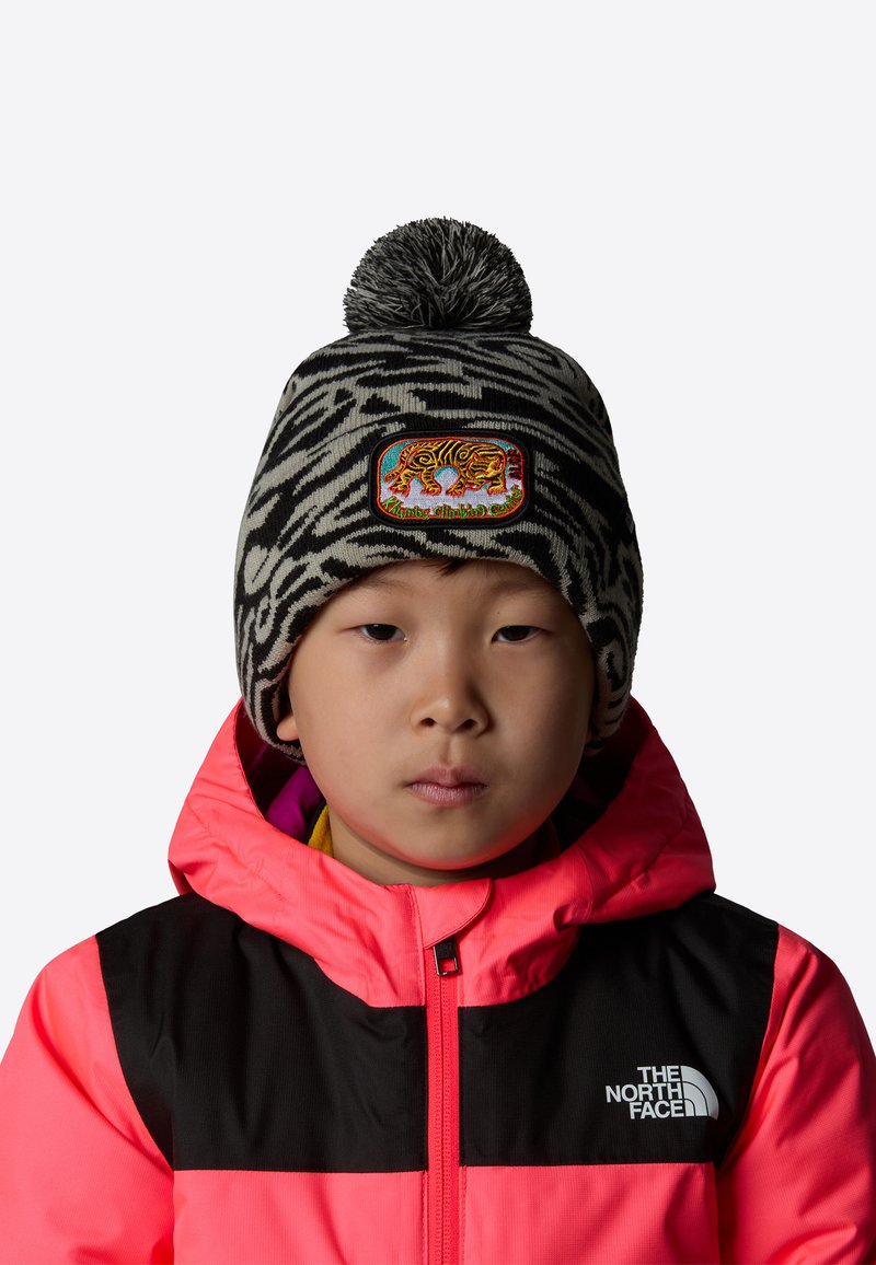 Czarna i biała czapka z dzianiny w wzór zebry, z kolorową naszywką tygrysa; pompon na górze. Noszona z jasnoróżową i czarną kurtką.