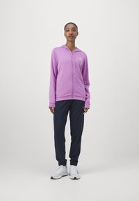 Fialová mikina na zips s kapucňou a bielym logom na hrudi, doplnená tmavomodrými teplákmi a bielymi športovými topánkami.