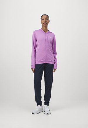Sudadera con cremallera morada, con capucha y logo blanco en el pecho, combinada con joggers azul oscuro y zapatillas deportivas blancas.