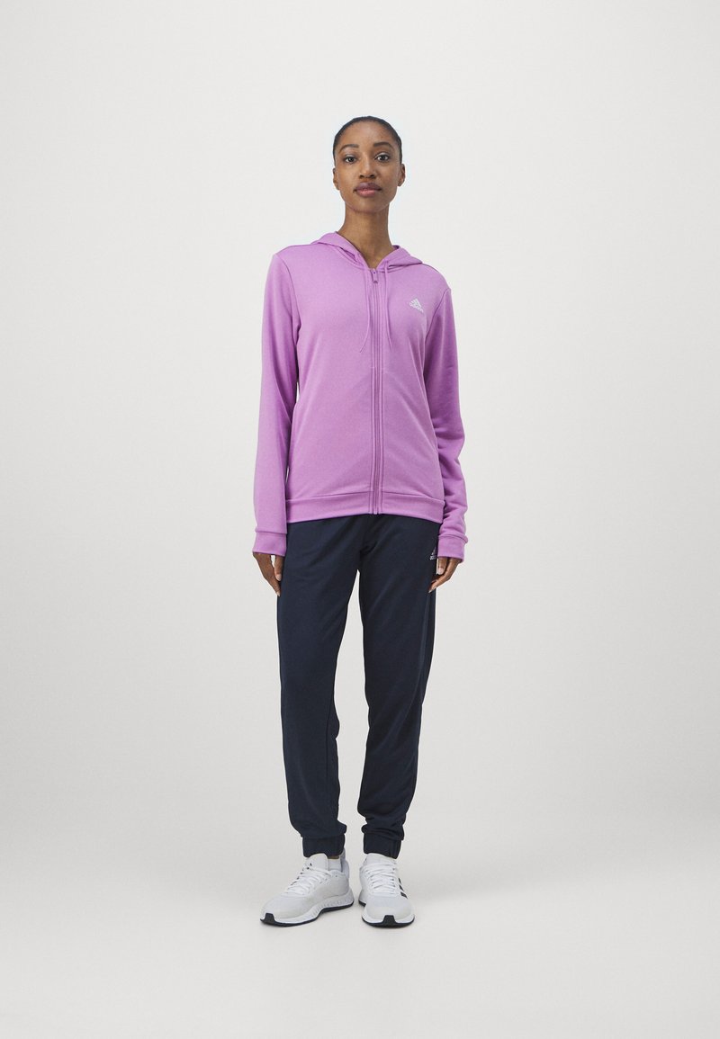 Fialová mikina na zips s kapucňou a bielym logom na hrudi, doplnená tmavomodrými teplákmi a bielymi športovými topánkami.