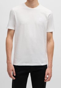 Bílý krátký rukáv tričko vyrobené z lehkého materiálu, s kulatým výstřihem a čtvercovou logovou nášivkou na levé hrudi.