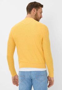 Gelber gestrickter Pullover mit langen Ärmeln, rundem Halsausschnitt und weicher Textur, kombiniert mit hellblauen Jeans und einem weißen Hemd darunter.