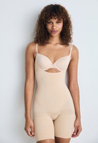 Beige shapewear bodysuit with adjustable straps, smooth texture, cut-out design at the bust, and mid-thigh length. Minimal seams and detailing.