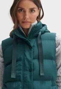 Türkisfarbene Steppweste mit Kapuze, ausgestattet mit silbernen Druckknöpfen, grünen Kordeln und einer gesteppten Textur. Getragen über einem hellgrauen Pullover.