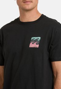 Mężczyzna noszący czarną koszulkę z małym prostokątnym graficznym wzorem niebieskich, czarnych i różowych fal oraz napisem "BILLABONG" na klatce piersiowej.