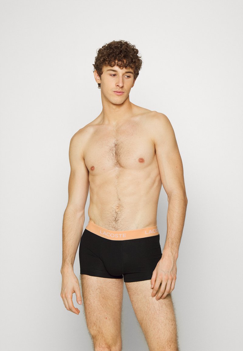 Zwarte katoenen trunks met een lichtoranje tailleband waarop het "Lacoste"-logo staat. Glad materiaal, aansluitend ontwerp, korte beenlengte.