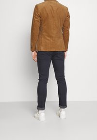 Hnědý kordový blazer s přiléhavým střihem, spárovaný s tmavě modrými slim-fit kalhotami a bílými teniskami; viditelné švy a ohrnuté rukávy.