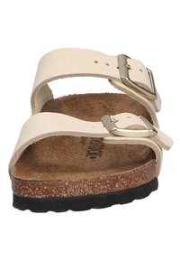 Beige sandals with two adjustable straps, metallic buckles, and a cork footbed. The sole is textured and black, providing a sturdy base.