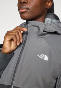 Grå vanntett jakke med glidelås foran, justerbar hette og "The North Face"-logo i hvitt på brystet.