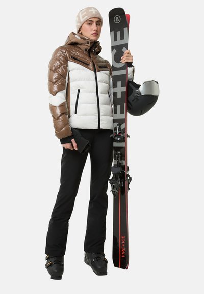 Gepolsterte braun-weiße Skijacke mit Reißverschlusstaschen; schwarze Skihose; schwarze Stiefel; hält schwarze Skier; trägt eine beige Mütze und Helm.