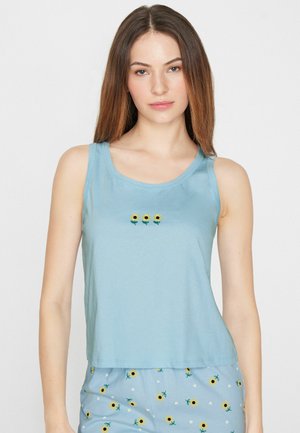 Jeune femme aux longs cheveux lisses porte un haut sans manches bleu clair avec trois petits motifs tournesols et un pantalon floral assorti.