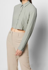 Camisa de manga larga cortada en tejido de color verde claro con textura, que presenta un cierre de botones en la parte delantera y un cuello extendido. Combinada con pantalones beige.