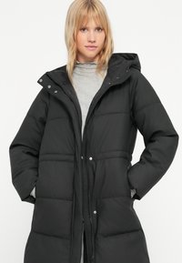 Čierny vypchávací dlhý kabát so zipsovým a patentkovým zapínaním, kapucňou a bočnými vreckami. Má hladkú textúru a prešívaný dizajn.