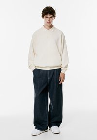 Pulover bej deschis cu guler, asortat cu pantaloni largi din denim închisi. Adidași albi cu o talpă texturată completează ținuta.