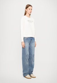 Felpa crema con logo 'Armani Exchange', abbinata a jeans a gamba larga azzurri chiari con grafiche 'AX' sulle gambe e sneaker beige.