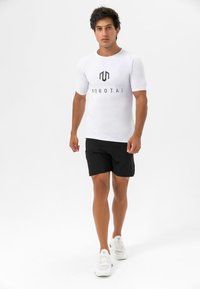 Hvit kortermet treningsskjorte med svart logo, kombinert med svarte shorts og hvite joggesko, stående mot en nøytral bakgrunn.