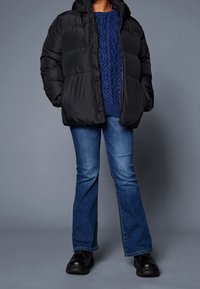Černá péřová bunda s kapucí, texturovaný modrý svetr s copánkovým vzorem, rozšířené modré džíny a černé boty. Pohodlný zimní outfit.