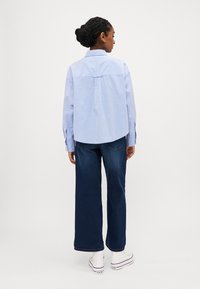 Mėlyna ilgomis rankovėmis marškiniai su kreipta apatine dalimi ir mygtukais susegamais rankogaliais, derinama su tamsiais plačiais džinsais ir baltais sportbačiais.