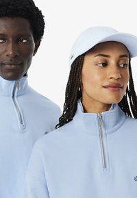 Dos personas con chaquetas azul claro con cremallera; una lleva una gorra a juego, ambas miran en direcciones ligeramente opuestas sobre un fondo simple.