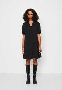 Černé šaty do délky kolen s krátkými nabíranými rukávy, véčkovým výstřihem a vrstveným spodním dílem. Oblékané s černými kozačkami po kolena a slunečními brýlemi.