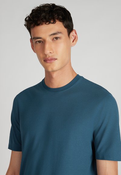 Joven con cabello corto y rizado, vistiendo una camiseta de cuello redondo en color teal, de frente contra un fondo gris claro liso.