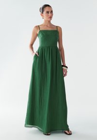 Garš zaļš kleita ar vertikālu tekstūru, kvadrātveida kakla izgriezumu, plānām siksnām un savelktu jostasvietu. Iespējo sānu kabatas un viļņotu apakšmalu.