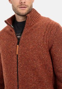 Rustfarbenes gestricktes Sweatshirt mit hohem geripptem Kragen und Reißverschluss, das ein strukturiertes Muster und subtile Farbtupfer aufweist.