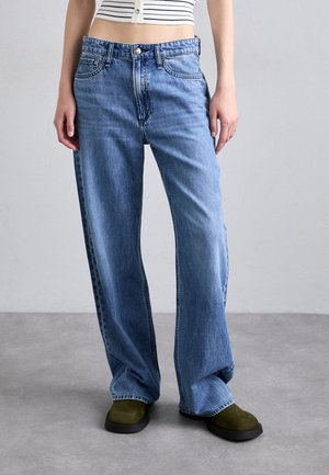 Kvinde iført højtaljede, vidde blå jeans og olivengrønne sko, stående på en grå overflade mod en ensfarvet lysegrå baggrund.