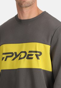 Grijze mesh lange mouw shirt met een dikke gele horizontale streep met "SPYDER" in zwarte letters op de borst. Textuurstof.