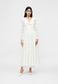 Γυναίκα με μακρύ λευκό φόρεμα από δαντέλα με βολάν στον γιακά και μακριά μανίκια, φοράει λευκά ψηλοτάκουνα παπούτσια με μυτερό μύτη και ιμάντα στο πίσω μέρος, στέκεται σε απλό φόντο.