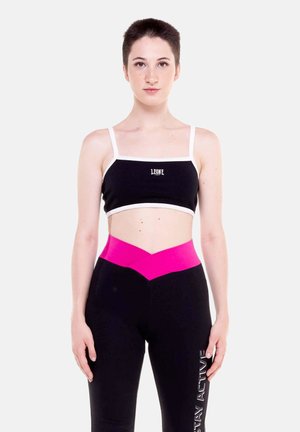 Jeune femme aux cheveux courts portant un crop top noir avec une bordure blanche et des leggings noirs avec une ceinture en V rose et un texte blanc sur la jambe.
